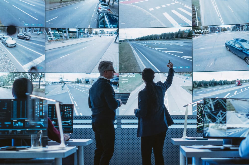 Two people in a control room analyze multiple screens displaying traffic camera footage of roads and intersections.