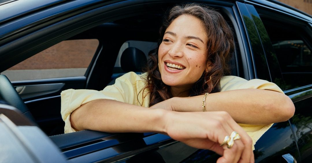 A woman smiling and leaning out of a car window, wearing a yellow shirt and jewelry, with a building in the background.
