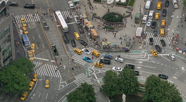 A bird's eye view of a busy intersection in New York City showing multiple modes of transport including buses, cars, taxis, bicycles, pedestrians etc.