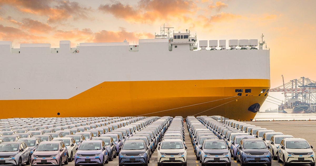A large cargo ship docked beside rows of parked cars at sunset.