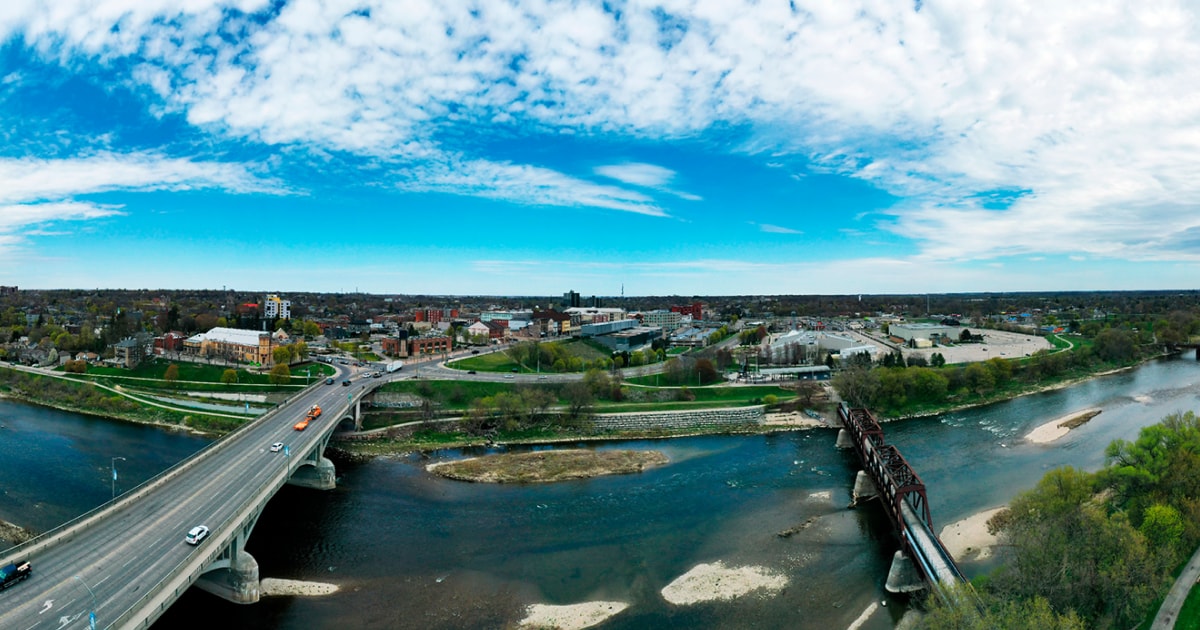 Aerial view of a cityscape with two bridges crossing a river, surrounded by green areas and buildings under a partly cloudy blue sky.