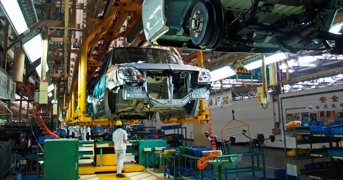 A partially assembled car is suspended from machinery in a factory, with workers and equipment visible in the background.
