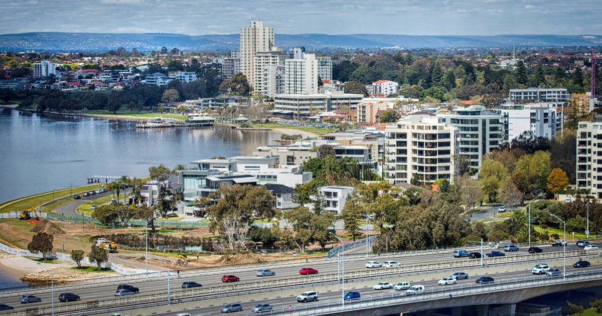Aerial view of a riverside urban landscape, featuring a mix of residential buildings, a highway with traffic, and green park areas along the water's edge.
