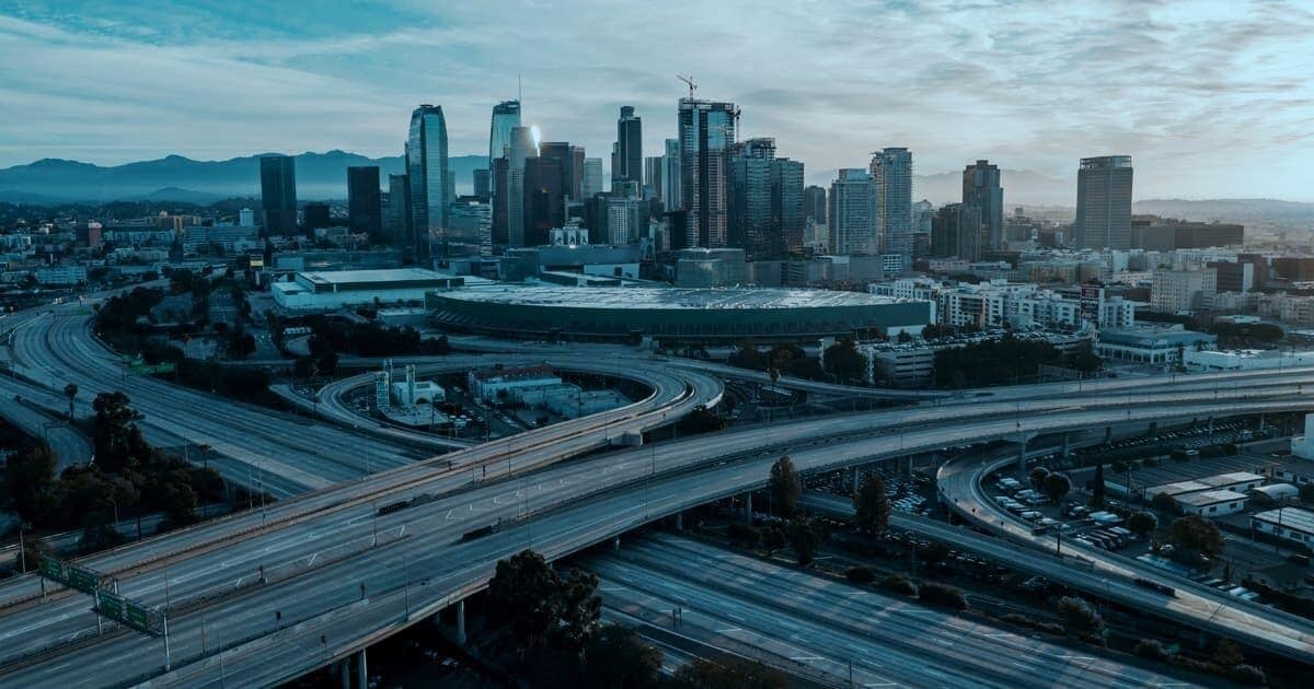 Aerial view of a city skyline with skyscrapers, intersecting highways, and surrounding urban landscape under a cloudy sky.