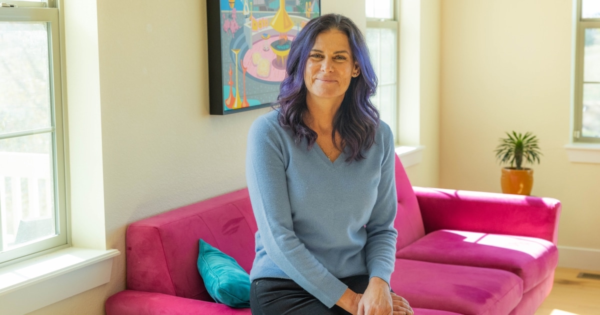 A woman with long wavy dark hair, highlighted with purple streaks, sits on a bright pink couch in a sunlit living room. She wears a light blue sweater and dark pants, smiling gently at the camera. Behind her is a colorful framed artwork, and a small potted plant sits near the window