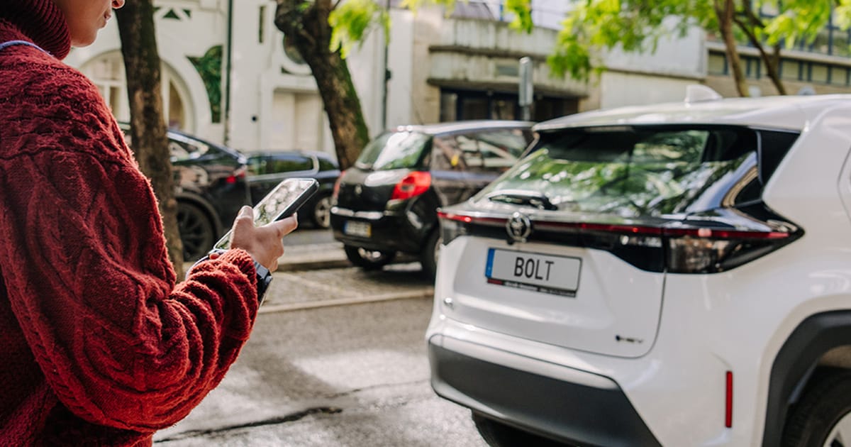 Person in a red sweater using a smartphone near a white car with a "BOLT" license plate, parked on a tree-lined street.