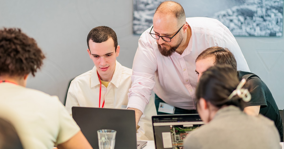 A bearded man in a pink shirt leans over to assist a group of four people working on laptops around a table. They appear focused, collaborating in a bright office environment.