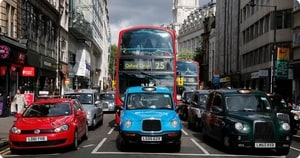 A head on picture of a typical road junction in London, showing cars, cabs, buses and pedestrians.