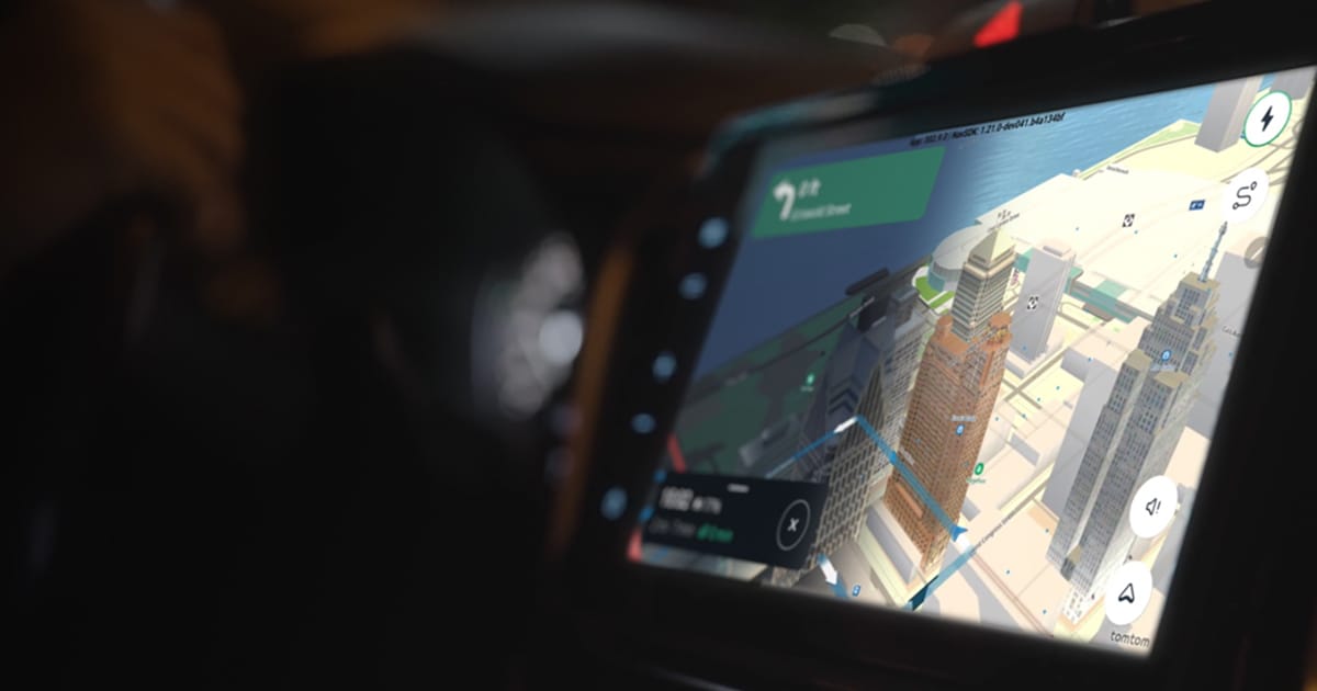 Car GPS screen displaying a 3D city map with directions. A hand is visible on the steering wheel in the dimly lit interior.