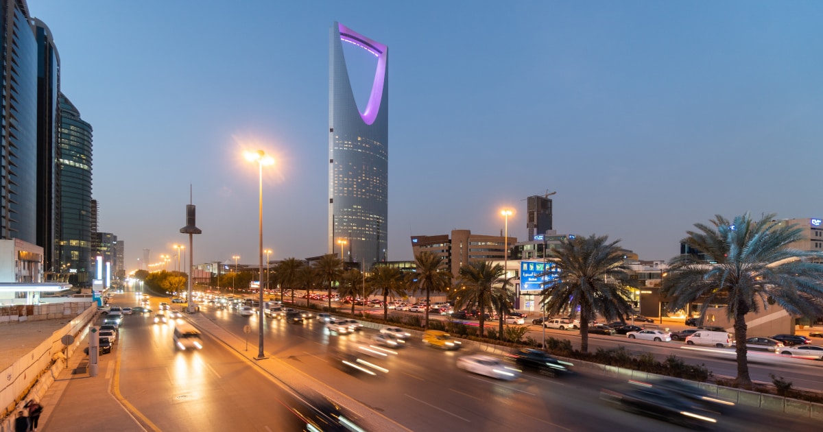City street in Riyadh at dusk, featuring the illuminated Kingdom Centre Tower and moving cars.