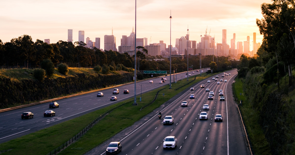 A sunset view of a busy highway with cars, framed by trees and the city skyline in the background.