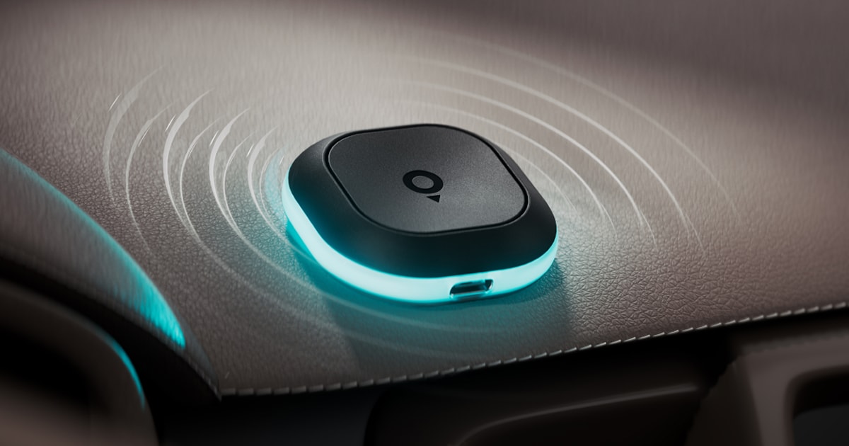 A small, black, circular device with a glowing blue edge rests on a car dashboard, emitting wave-like signals.