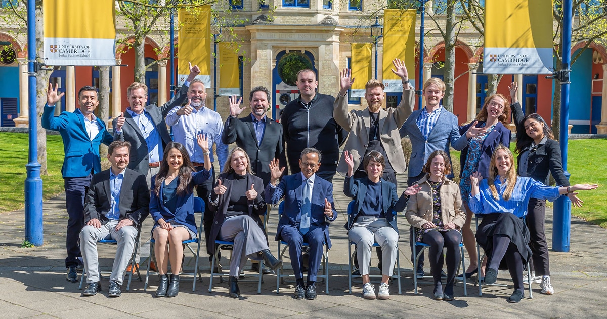 A diverse group of professionals posing cheerfully in front of the University of Cambridge Judge Business School, under yellow banners. Some are seated while others stand behind them, raising their hands in a celebratory gesture