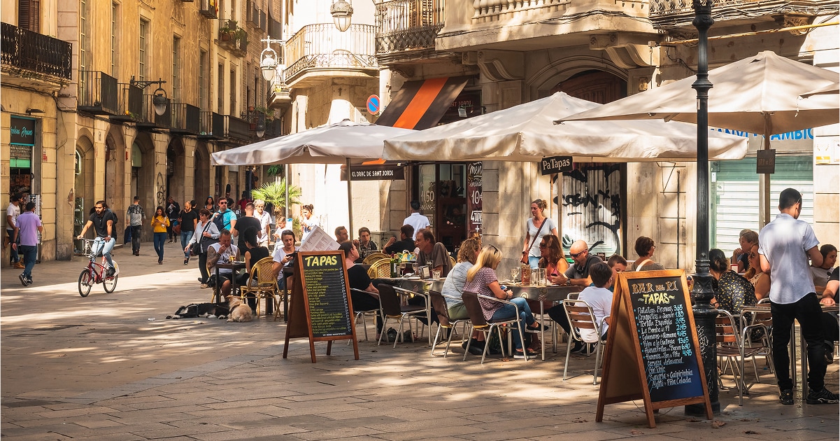 Outdoor café with people dining under umbrellas on a sunny street. Chalkboard menus display "Tapas." A cyclist and pedestrians pass by.