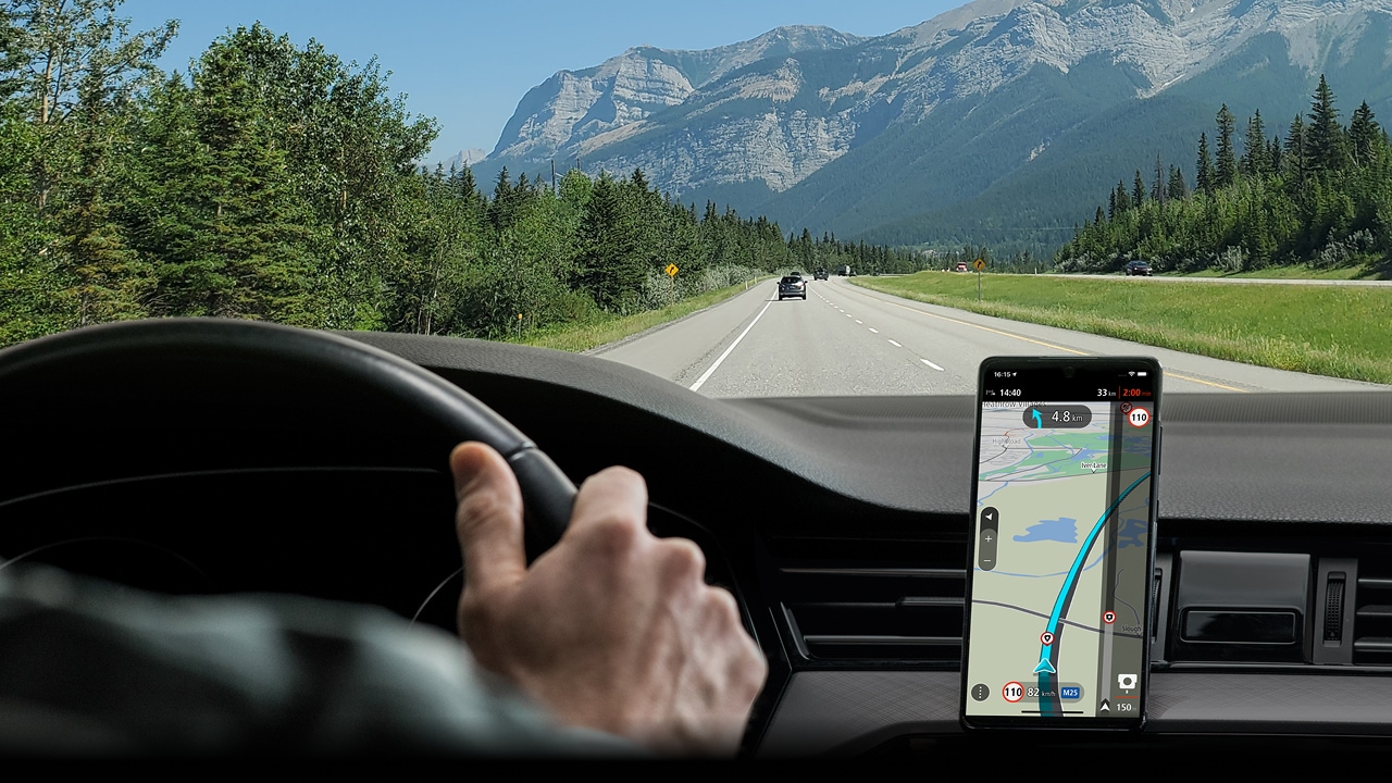 Driver's view of a scenic mountain road with a GPS navigation app on a smartphone mounted on the dashboard.