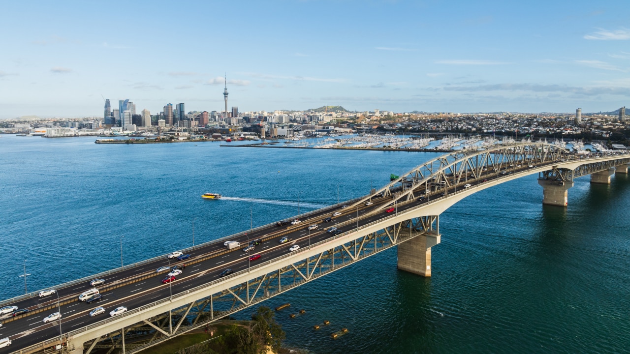 Aerial view of a bridge with vehicles crossing over water, leading to a city skyline featuring tall buildings and the Sky Tower in Auckland, New Zealand.
