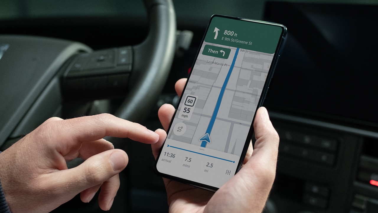 Close-up of hands holding a smartphone with a navigation app displayed, showing a route and speed limit, inside a car.
