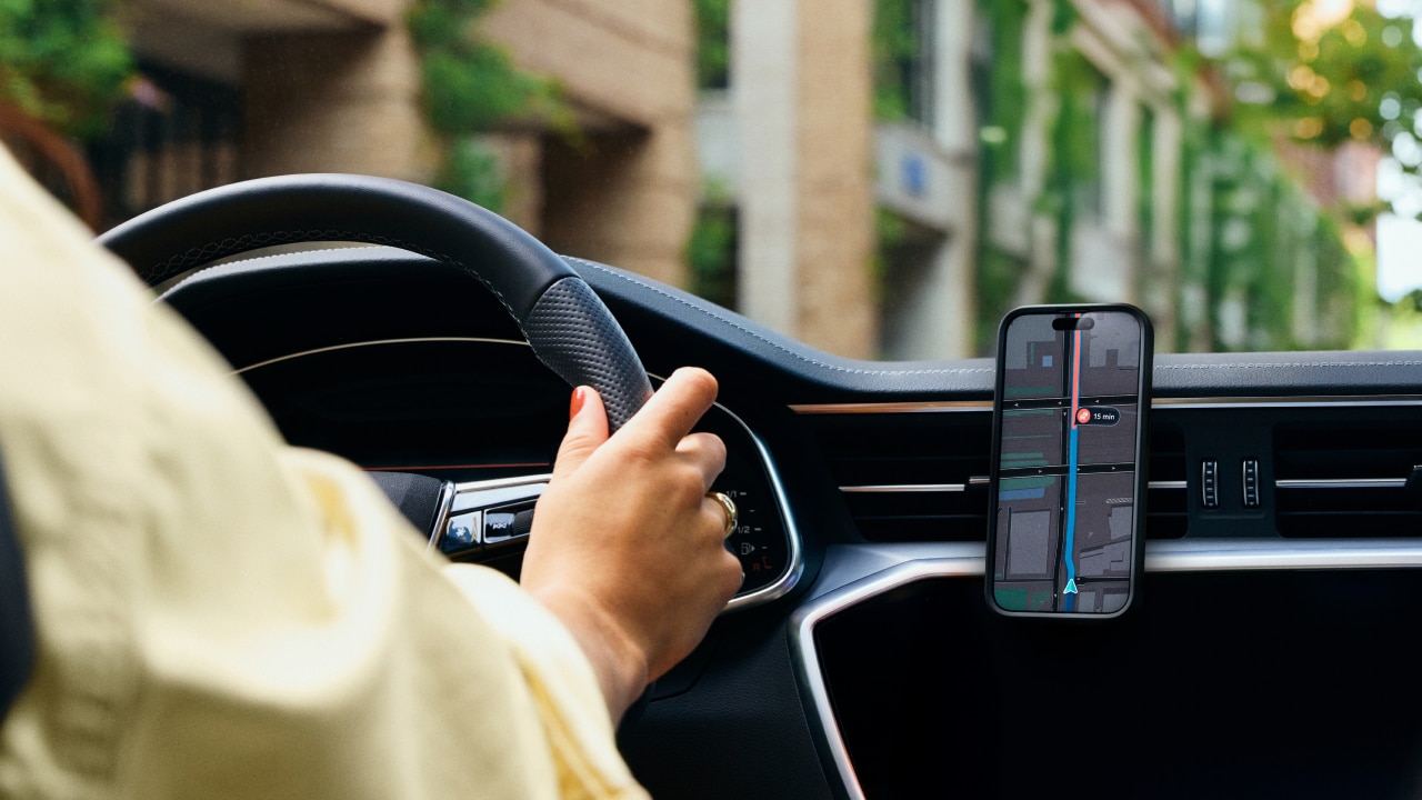 Person driving a car, holding the steering wheel with one hand. Smartphone with GPS navigation is mounted on the dashboard.