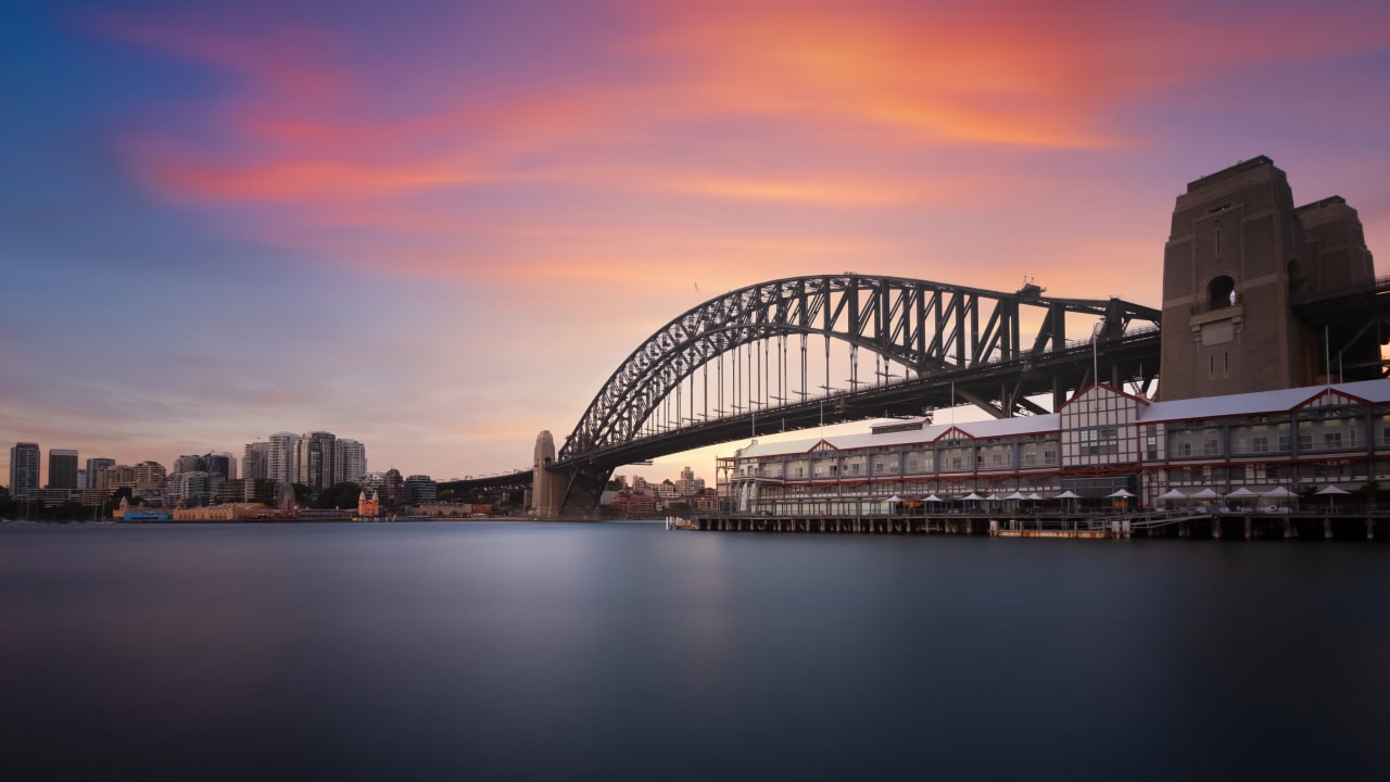 A sunset view of the Sydney Harbour Bridge, with colorful skies and a waterfront building in silhouette.