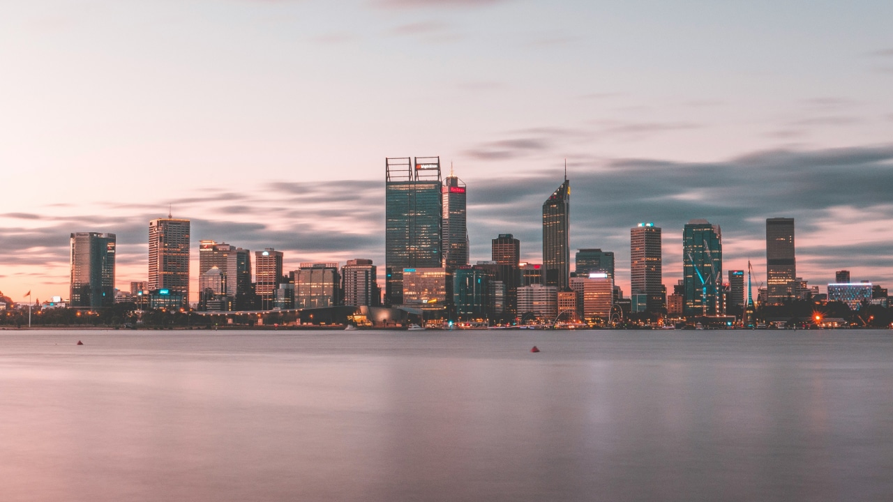 A panoramic view of a modern skyline at dusk, featuring tall skyscrapers reflecting soft evening light over a calm body of water.