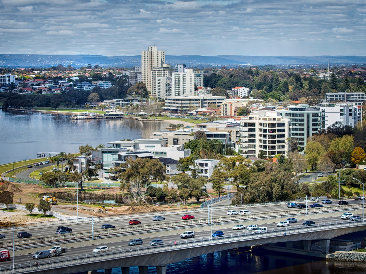 Aerial view of a riverside urban landscape, featuring a mix of residential buildings, a highway with traffic, and green park areas along the water's edge.