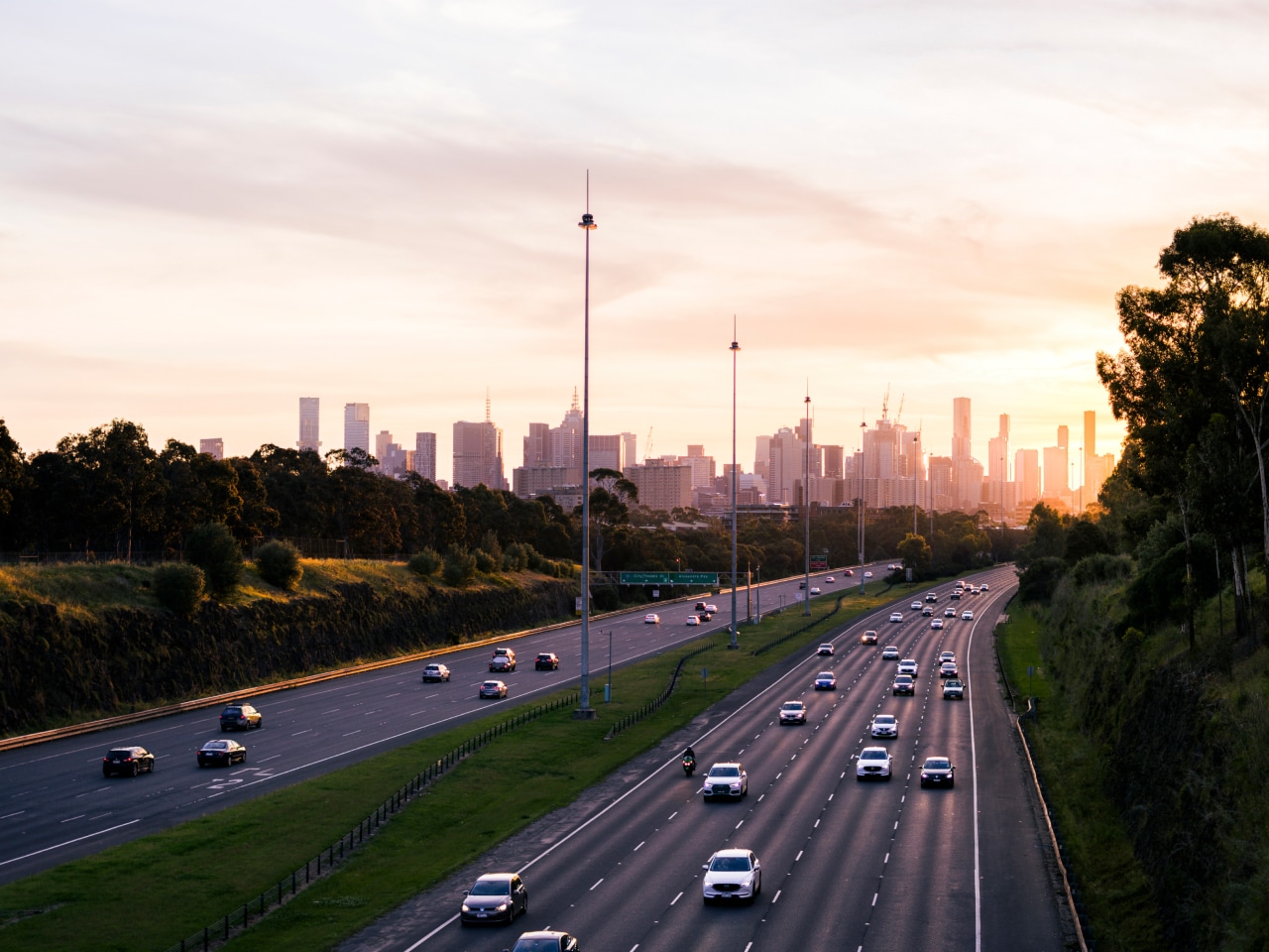 A sunset view of a busy highway with cars, framed by trees and the city skyline in the background.