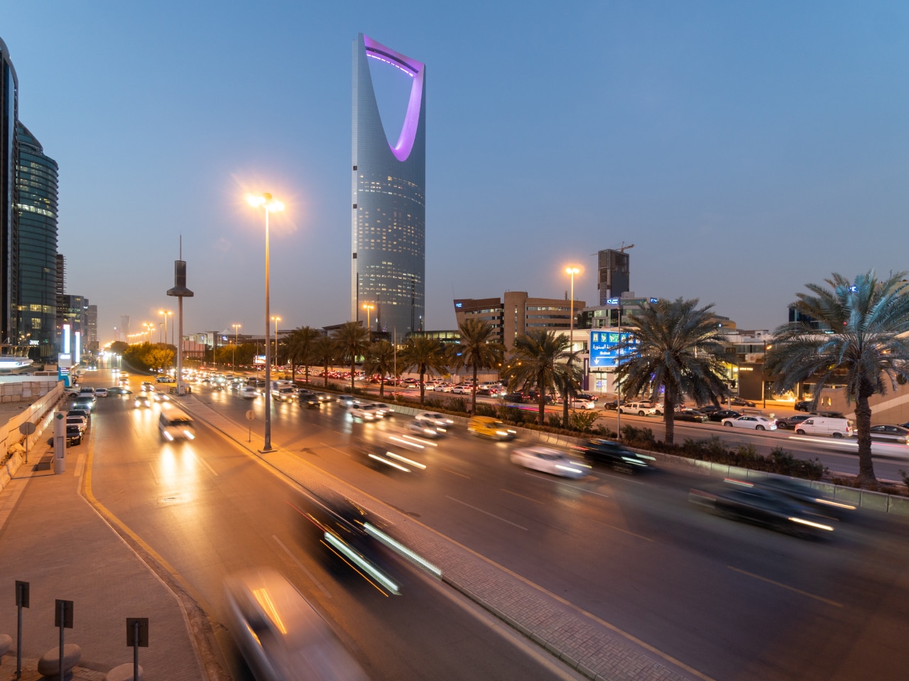 City street in Riyadh at dusk, featuring the illuminated Kingdom Centre Tower and moving cars.