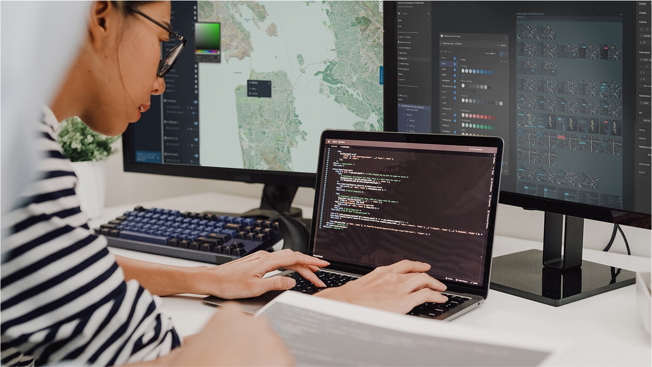 Person working on a laptop with code on the screen, surrounded by two large monitors displaying maps and data visualizations.