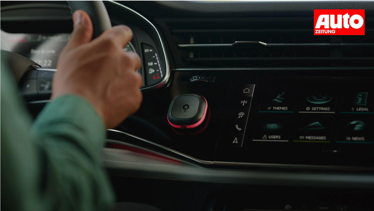 A person holds a steering wheel beside a car dashboard with a touchscreen panel displaying various icons and a mounted device.