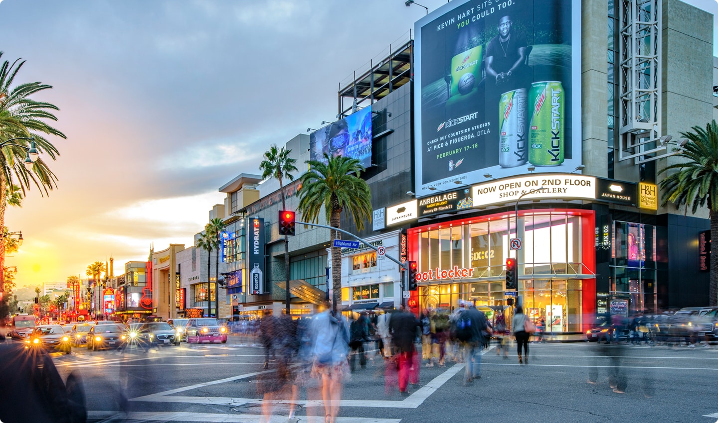Busy city street at sunset with blurred pedestrians, palm trees, and illuminated storefronts, including a large Foot Locker and billboard ads.