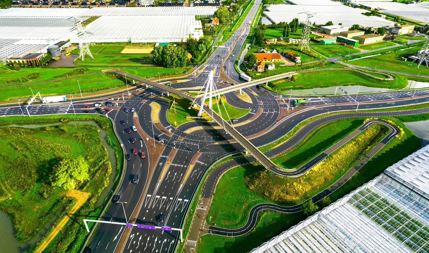 Aerial view of a complex road interchange with multiple lanes and overpasses, surrounded by green fields and industrial buildings.