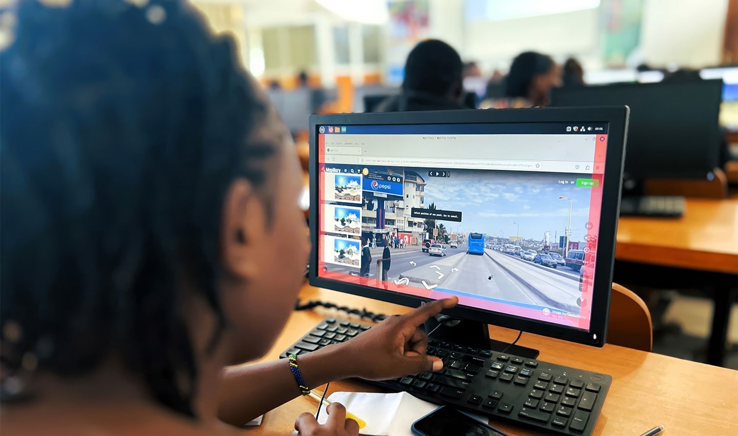 A person pointing at a computer screen displaying a street view, set in a busy computer lab.