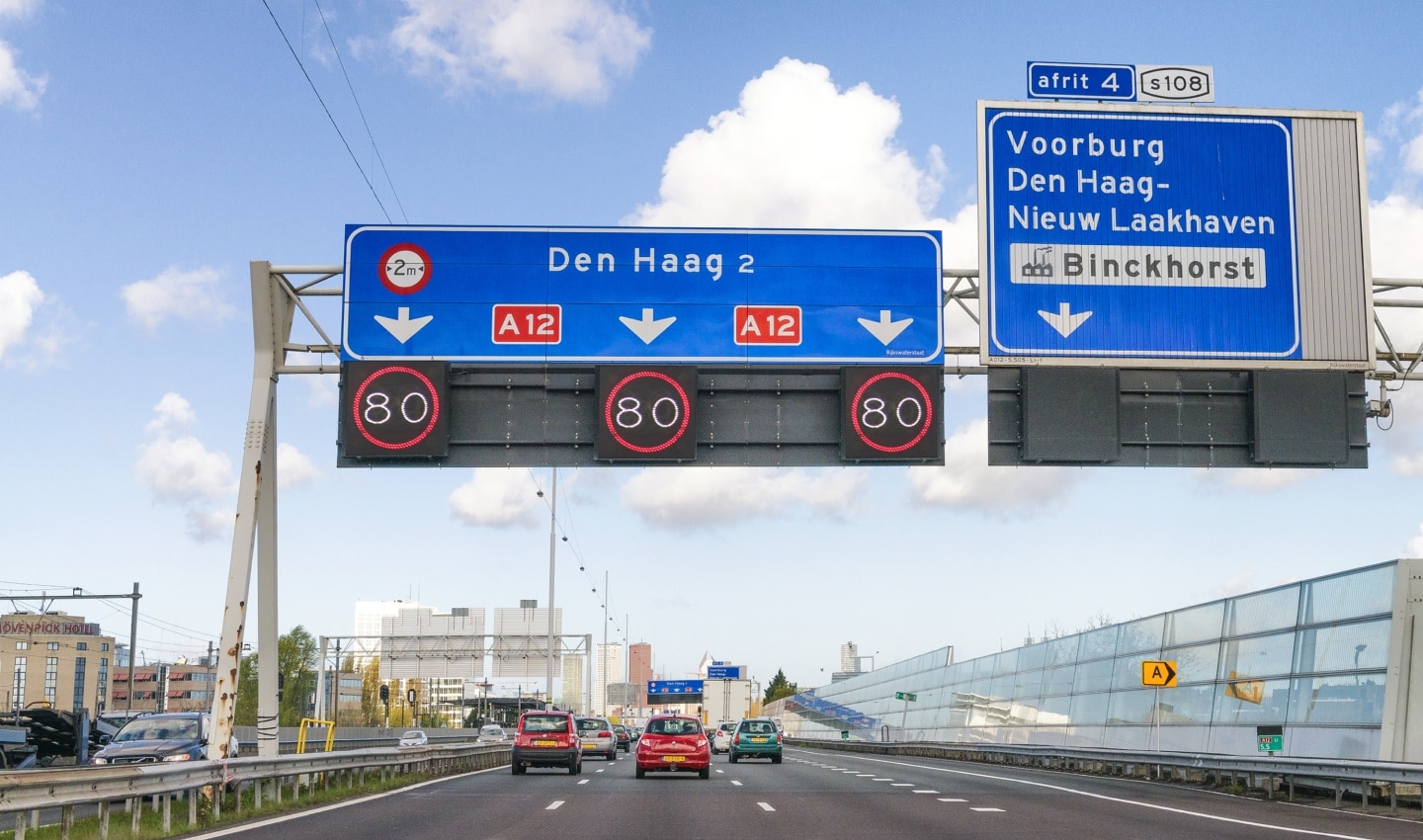 Highway view with cars and overhead signs showing directions to Den Haag and Voorburg on A12, speed limit 80 km/h. Clear sky and city skyline ahead.