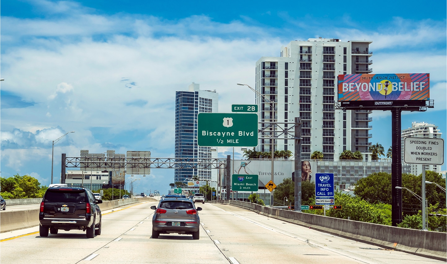 Highway scene with cars, Biscayne Blvd exit sign, tall buildings, and billboards under a blue sky with clouds.