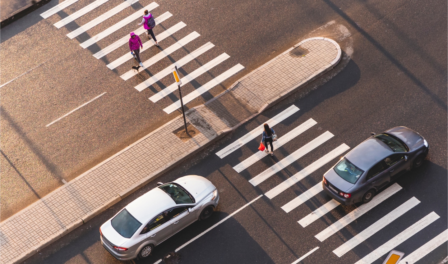 An aerial view of a crosswalk with two pedestrians walking, while cars approach the intersection, highlighting the urban setting.