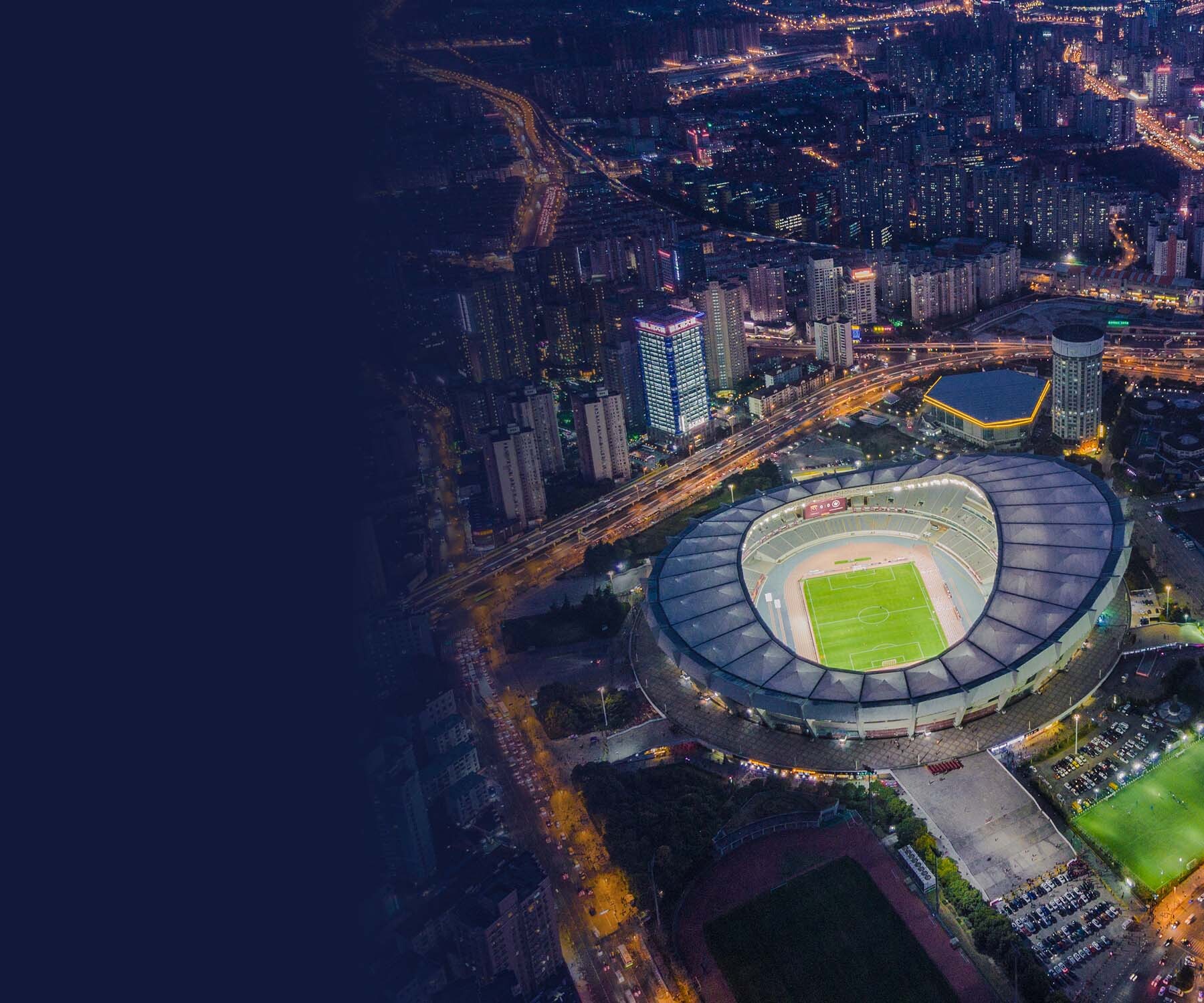 Aerial view of a brightly lit sports stadium in a cityscape at night, surrounded by illuminated buildings and roads.