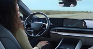 A person sits in a modern car with digital dashboard displays, showing speed and GPS navigation, driving on an open road.