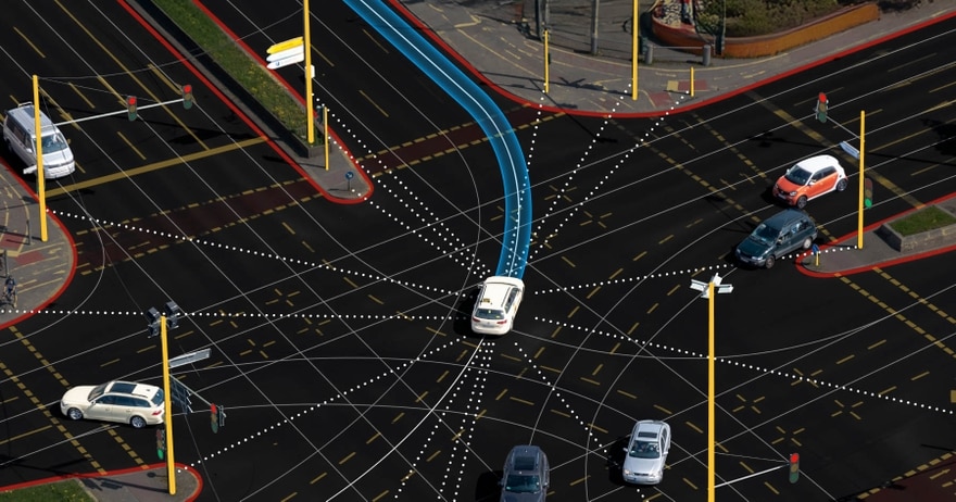 Aerial view of a city intersection with multiple cars, traffic lights, and digital navigation paths indicated by lines and dots.