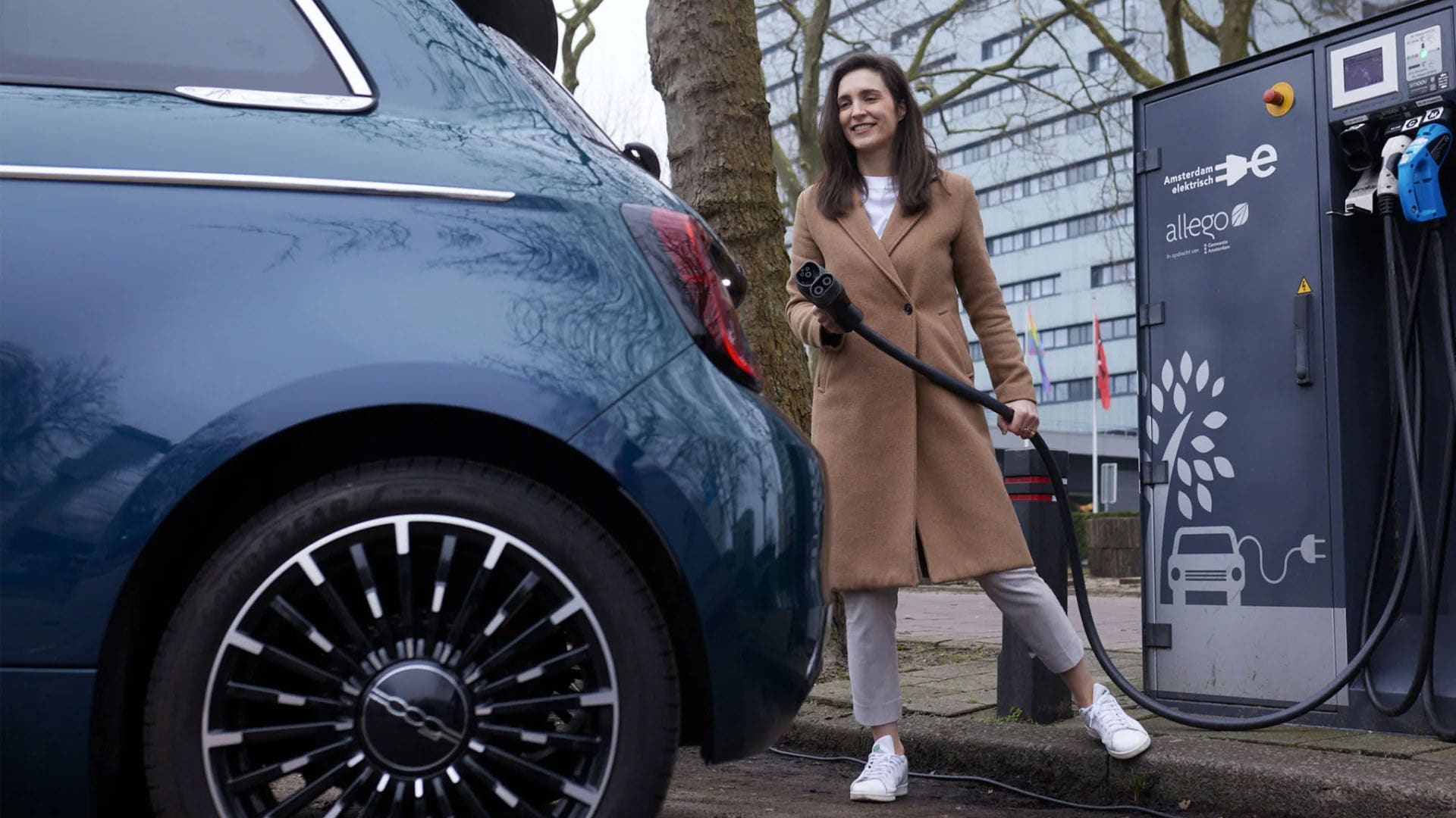Woman in a beige coat holds a charging cable near a blue electric car at a charging station, with trees and a building in the background.