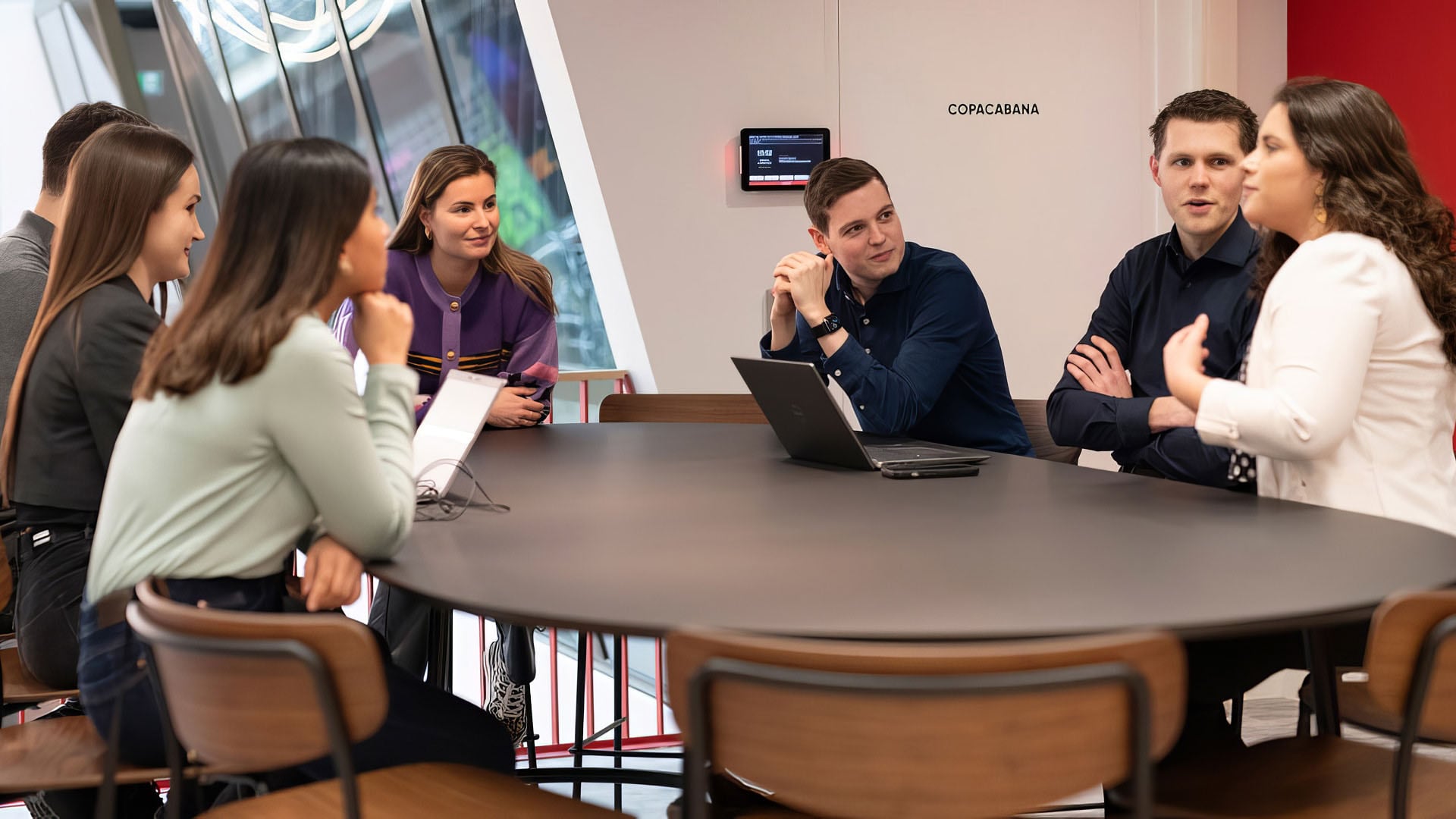 A group of people engaged in a meeting around a round table, some using laptops, in a modern conference room setting.