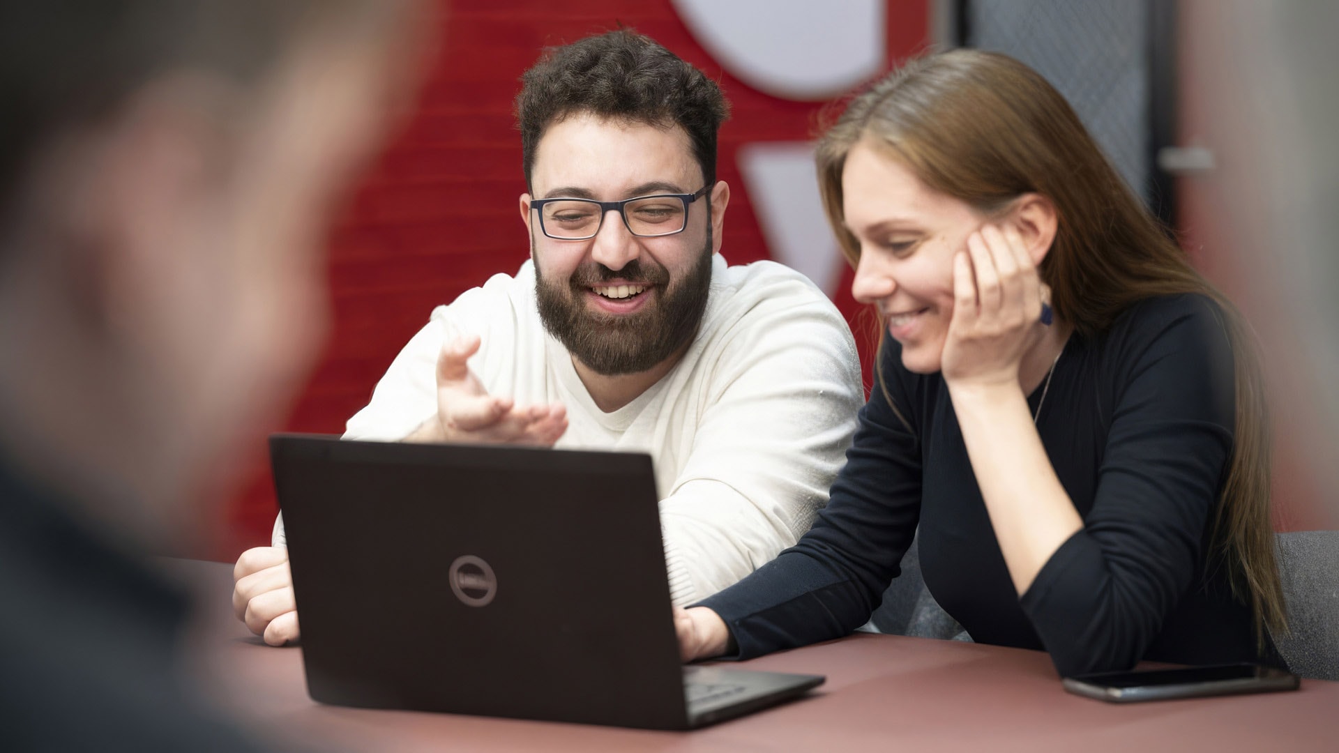 A group of people engaged in a meeting around a round table, some using laptops, in a modern conference room setting.