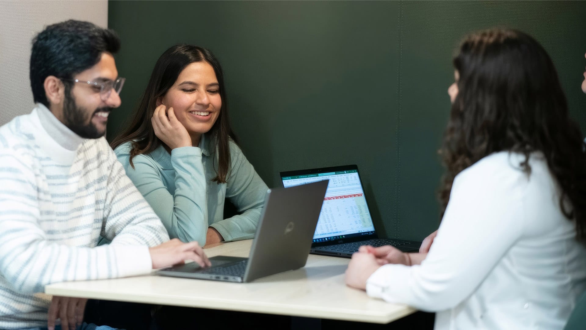 A group of people engaged in a meeting around a round table, some using laptops, in a modern conference room setting.