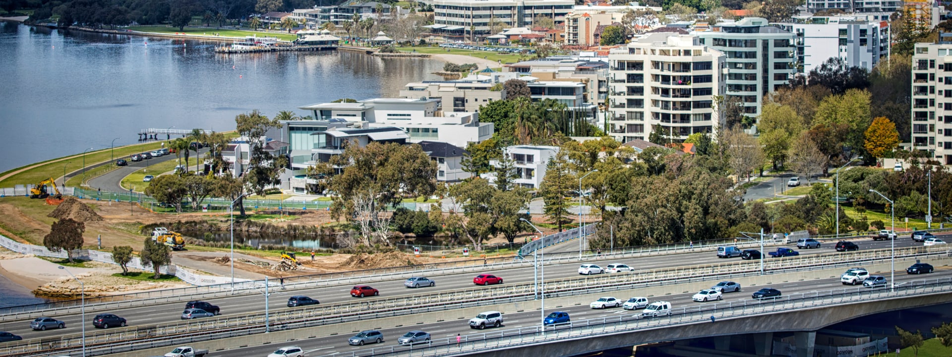 Aerial view of a riverside urban landscape, featuring a mix of residential buildings, a highway with traffic, and green park areas along the water's edge.