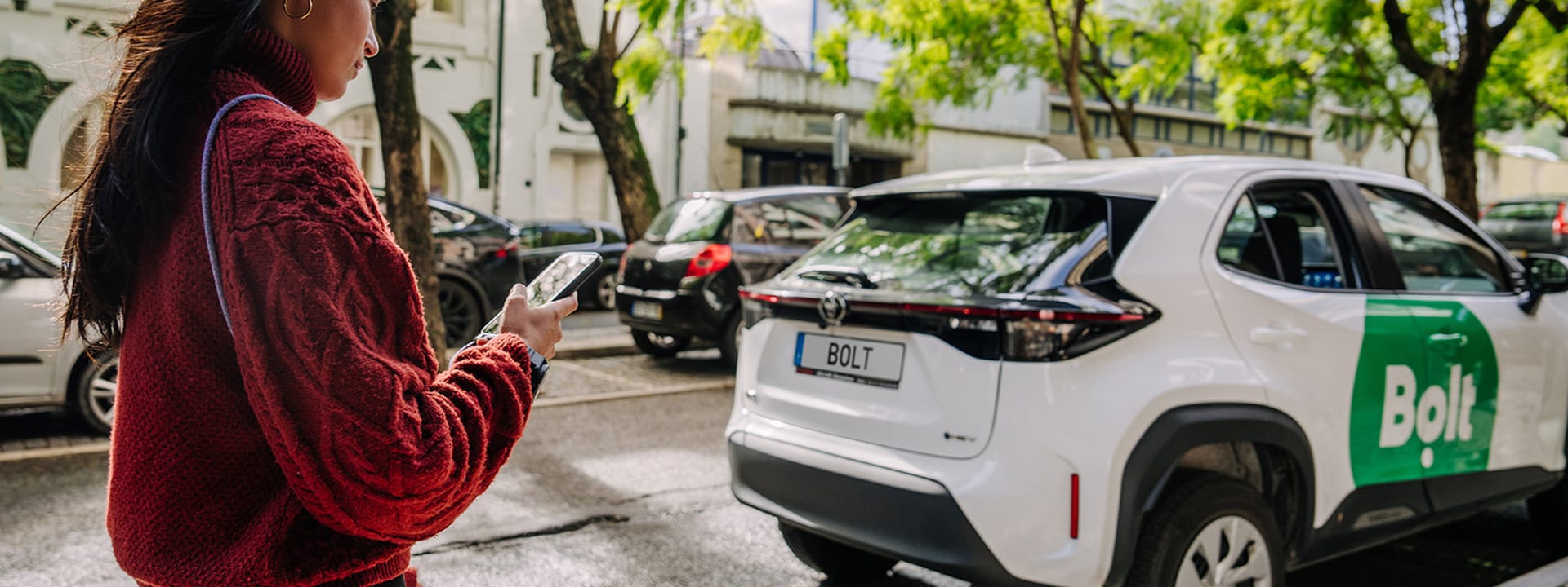 Person in a red jacket using a smartphone stands near a Bolt ride-sharing car parked on a tree-lined street.