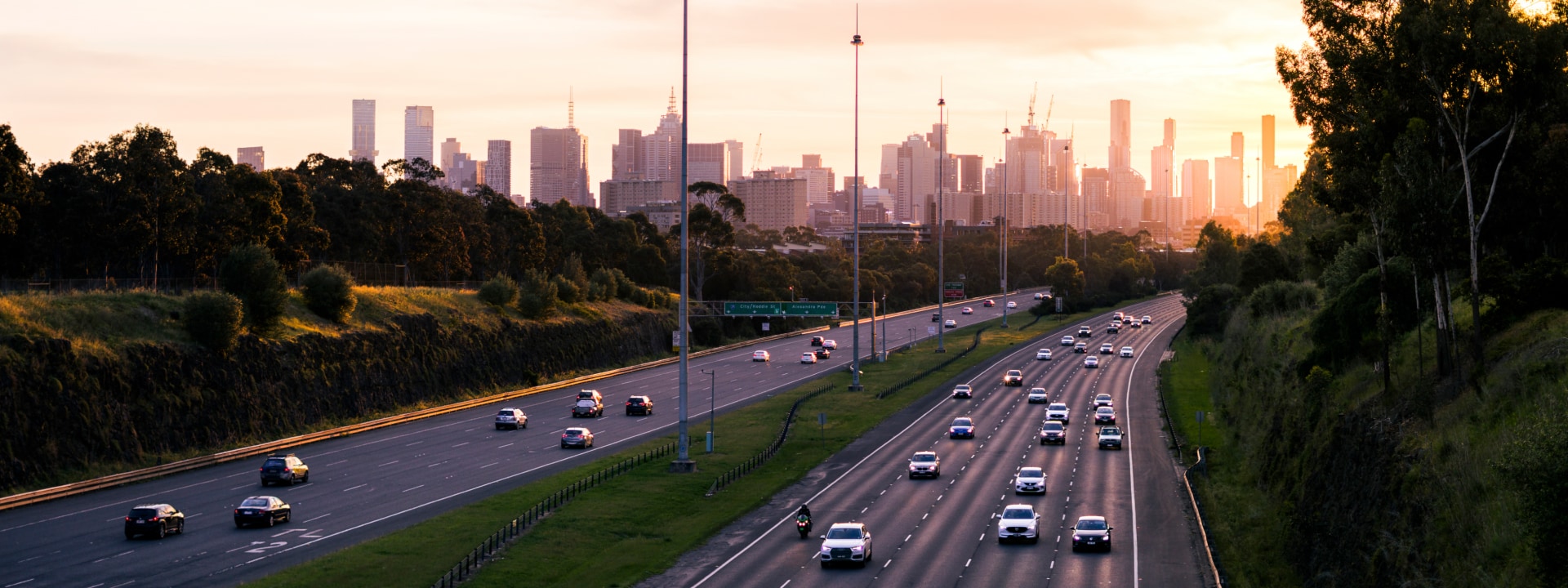 A sunset view of a busy highway with cars, framed by trees and the city skyline in the background.