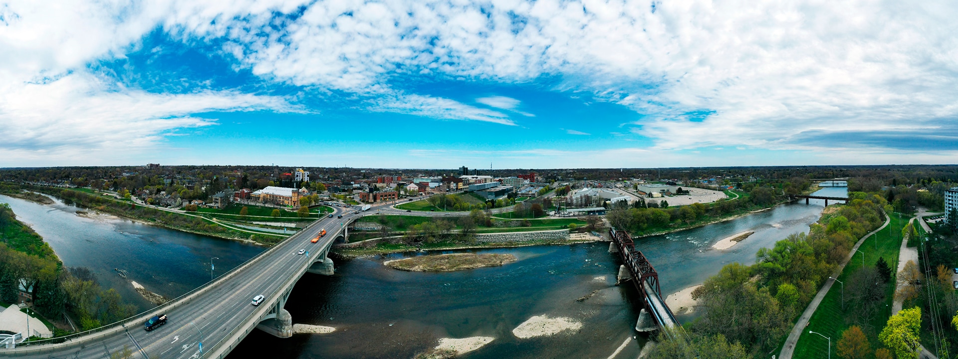 Aerial view of a cityscape with a river, bridges, and roads under a partly cloudy sky.