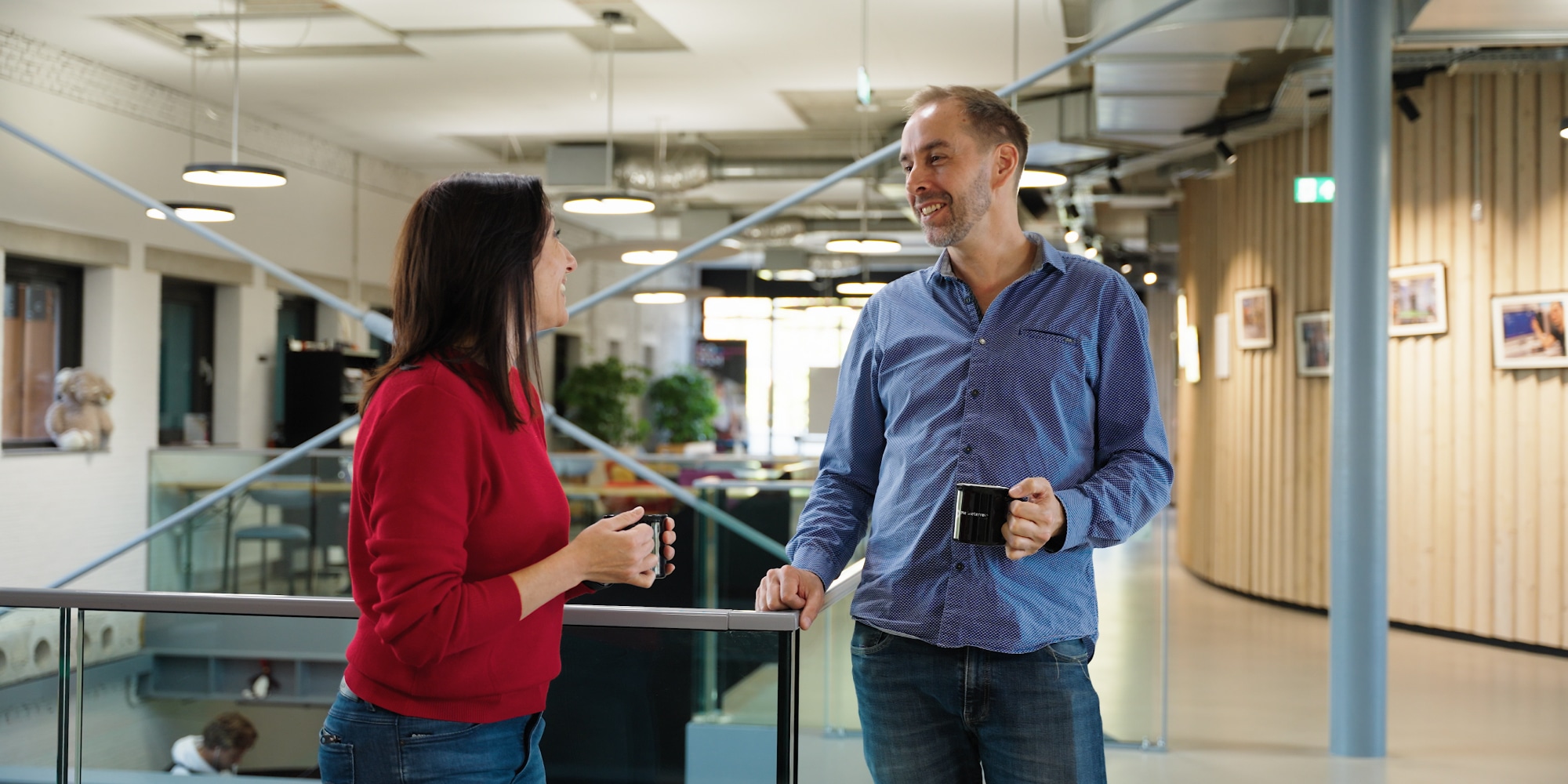 A woman in a red sweater and a man in a blue shirt chat while holding mugs in a modern office hallway.