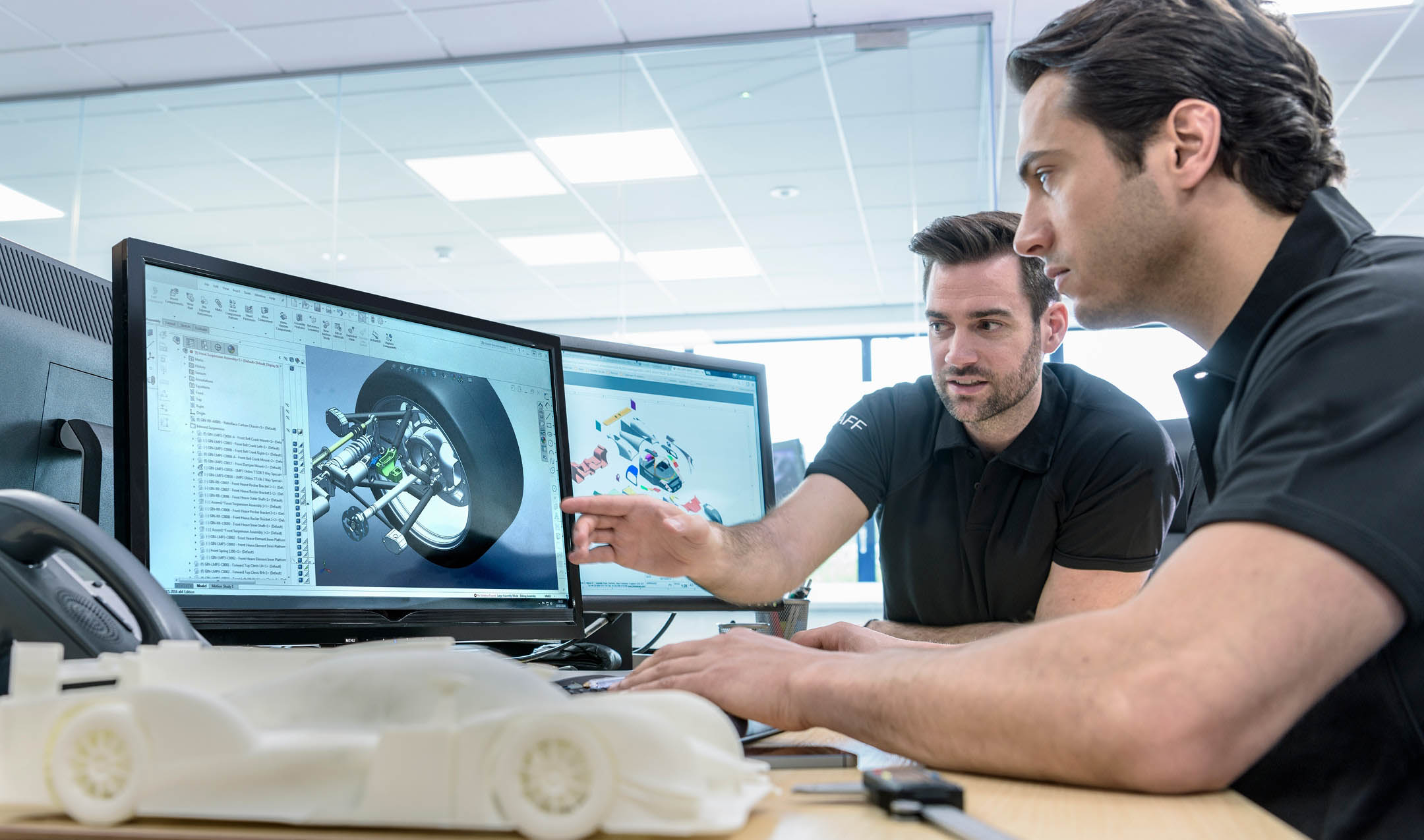 Two carmakers discuss a design displayed on dual computer monitors.
