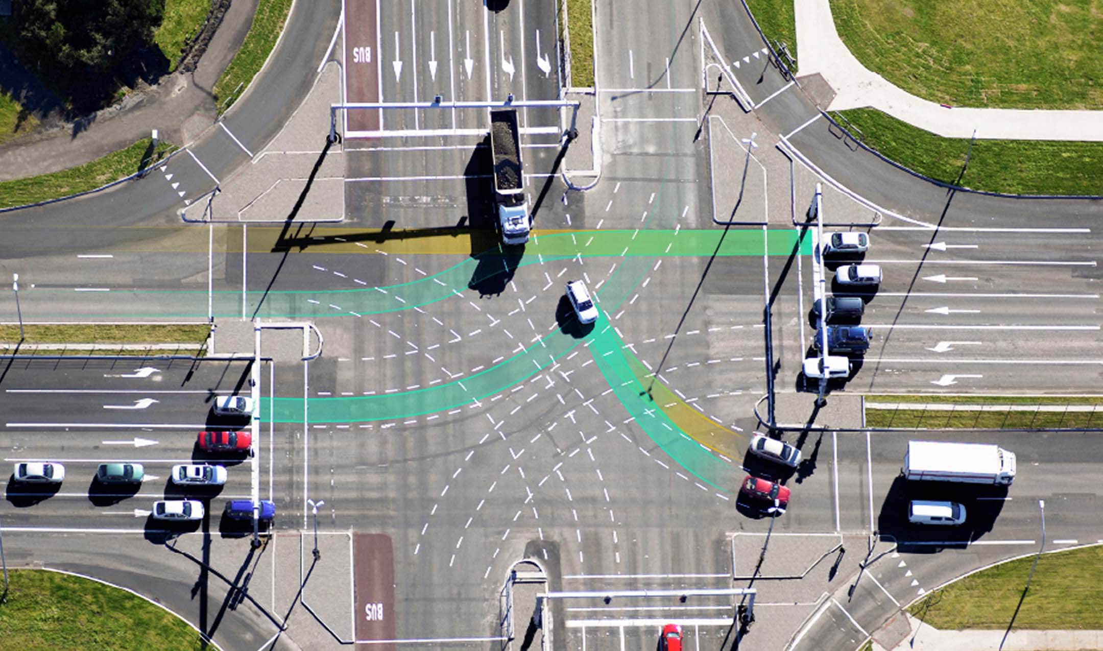 Aerial view of a busy intersection with multiple lanes, vehicles, traffic markings, and highlighted pathways, surrounded by grass and sidewalk areas.