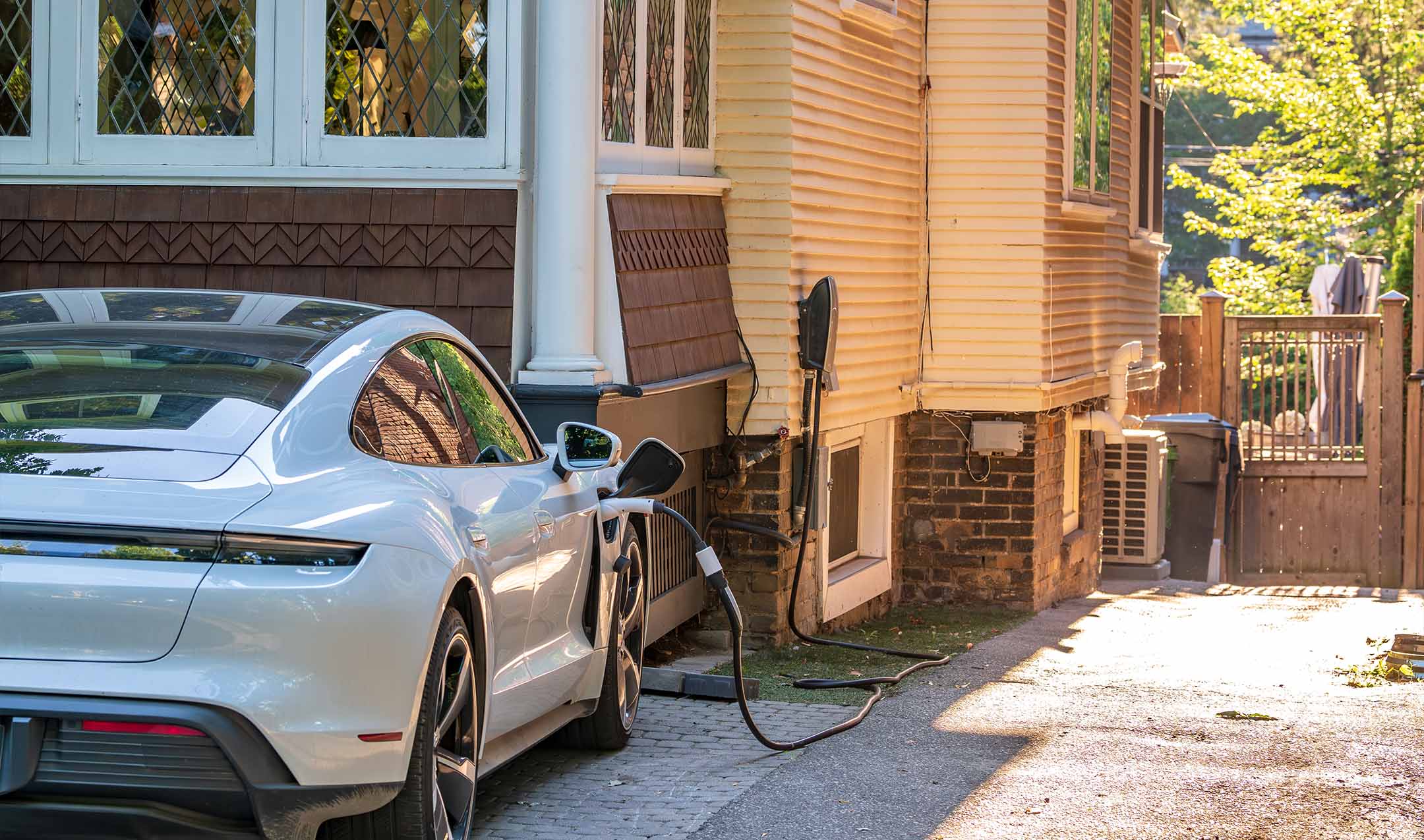 A white electric car is parked in a driveway, plugged into a charging station mounted on a house wall on a sunny day.
